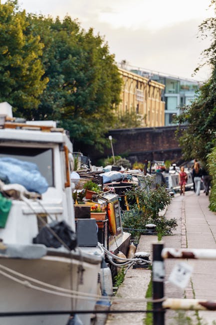 The image depicts a narrow urban canal-side pathway with a mix of boats moored along the water's edge, some covered with tarps and others exposed, revealing details like ropes, fenders, and equipment.