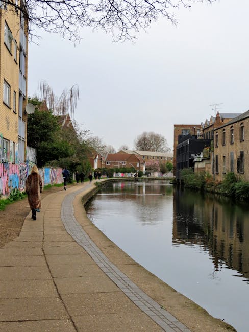 A paved pathway alongside a narrow canal in a residential area, with various plants and small trees lining the edge. To the left, there are multi-storey buildings with some graffiti on the lower walls